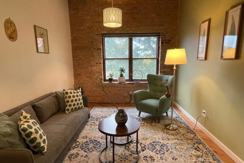 A look at one of the rooms in the Chicago Minds coworking space. It has an exposed brick wall with a window lined with potted plants. The room is designed in hues of green, grey, blue, and brown. There is a green chair with a reading lamp opposite a grey couch with toss cushions, including a polka dot white and green pillow. A table with a vase sits in the center and underneath that is an area rug. A pendant light hangs from the ceiling. There are framed pictures on the wall of different leaves and foliage.