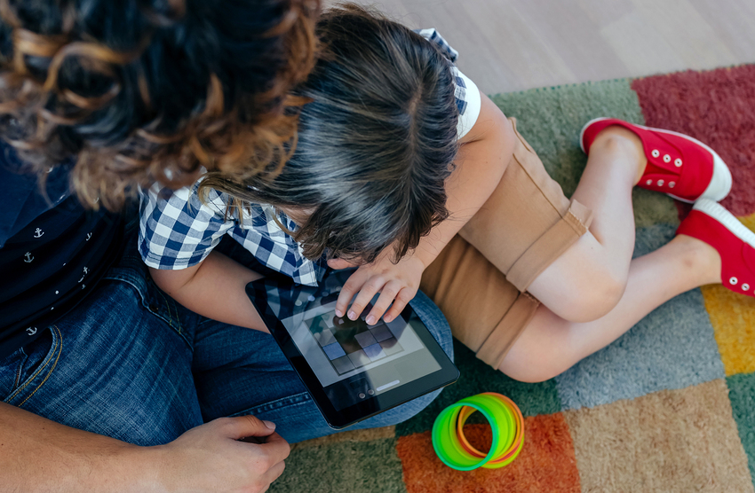A child and their parents play a game on a tablet together while sitting on a playmat in a waiting room