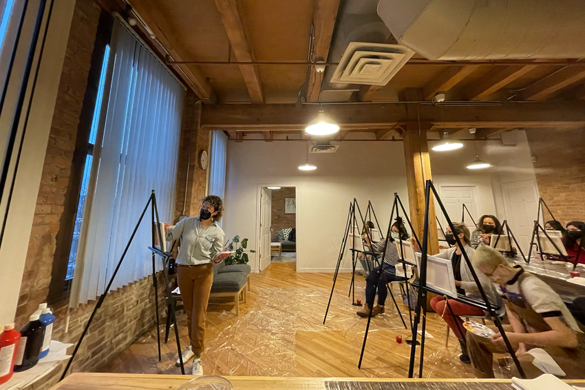 A paint night hosted in the Chicago Minds space. There are two rows of community members with easels, painting on canvases. Someone leads the group at the front of the room with their own canvas on an easel. The room is large with high ceiling and an exposed brick wall at the front with windows that have closed blinds.