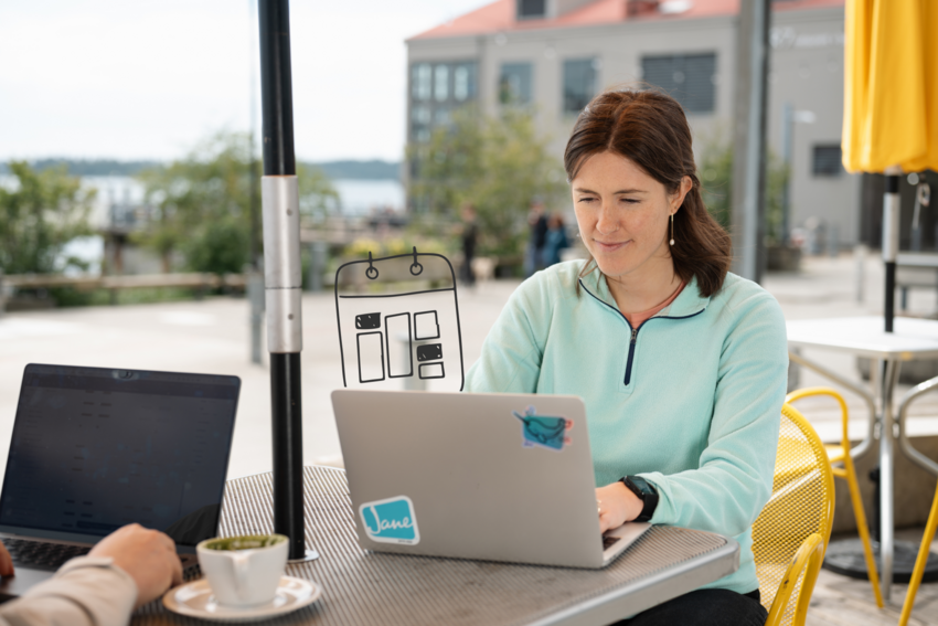 Woman working on a laptop with a Jane sticker at an outdoor café table, coffee nearby