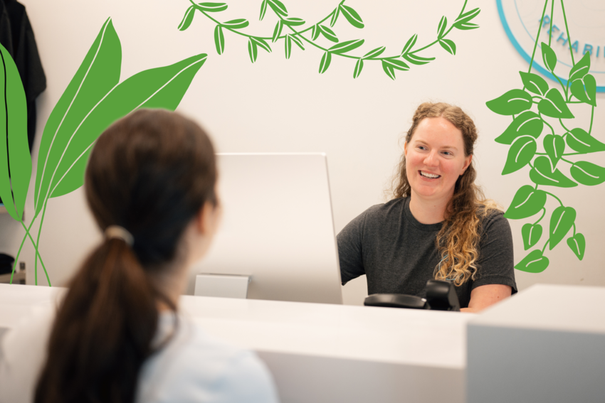 A woman behind a front desk smiles at a patient. Three illustrations of plants surround her, including a fern on the floor and hanging plants.