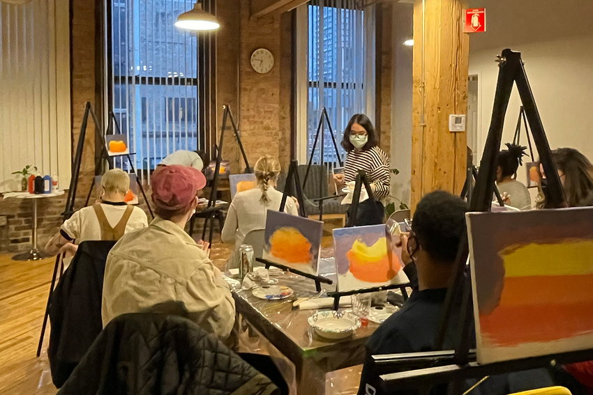Another view of paint night in the Chicago Minds space, this time from the perspective of the painters. A diverse group all paints the same image on their canvas: a swirl of red, yellow, and orange against a blue backdrop.