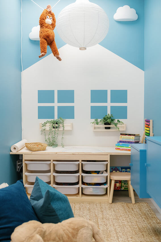 A waiting room designed with storage bins with the backdrop of a house, clouds, and potted planters. A stuffed monkey hangs from the ceiling attached to an overhanging pendant light.