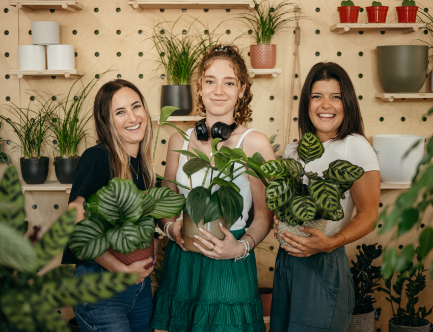 Sonia, Julie, and Jacquie of Plantsome smile for the camera with plants in their arms against a plant wall.