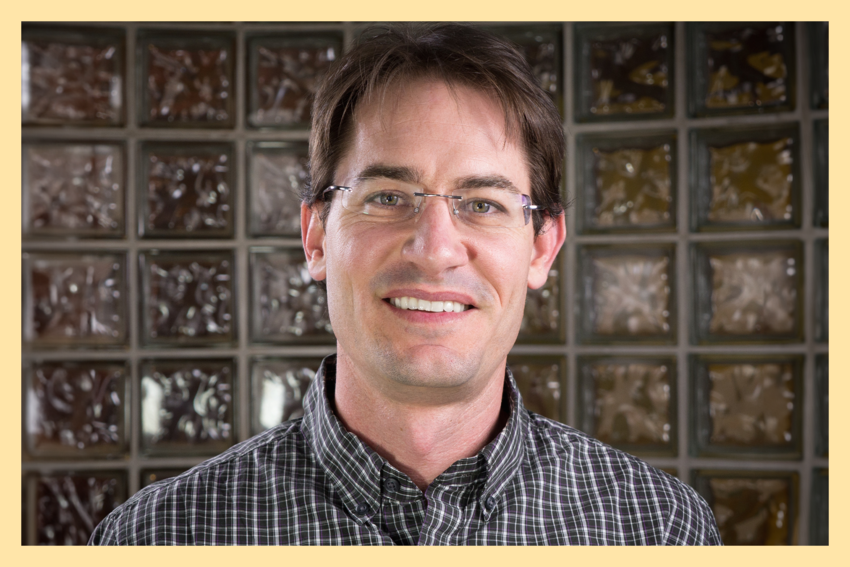Travis Good, CEO of HIPAAYak, smiles for the camera. He wears a red and white checkered shirt and glasses, and stands against a backdrop of a frosted glass wall. There is a light yellow border around his photo.