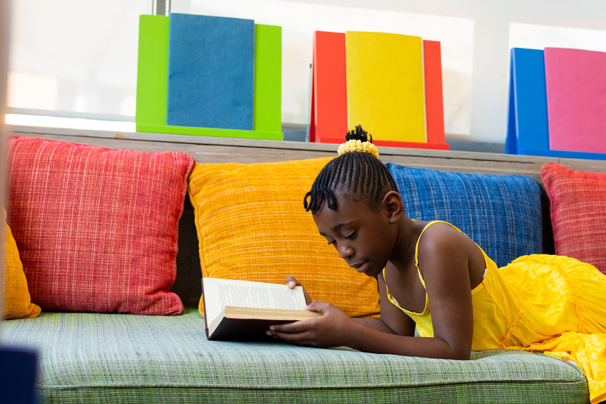 A young girl lies down on a couch in a waiting room and reads a book