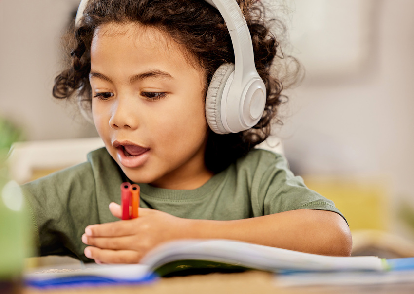 A young child with headphones on holds markers as they look at an activity book